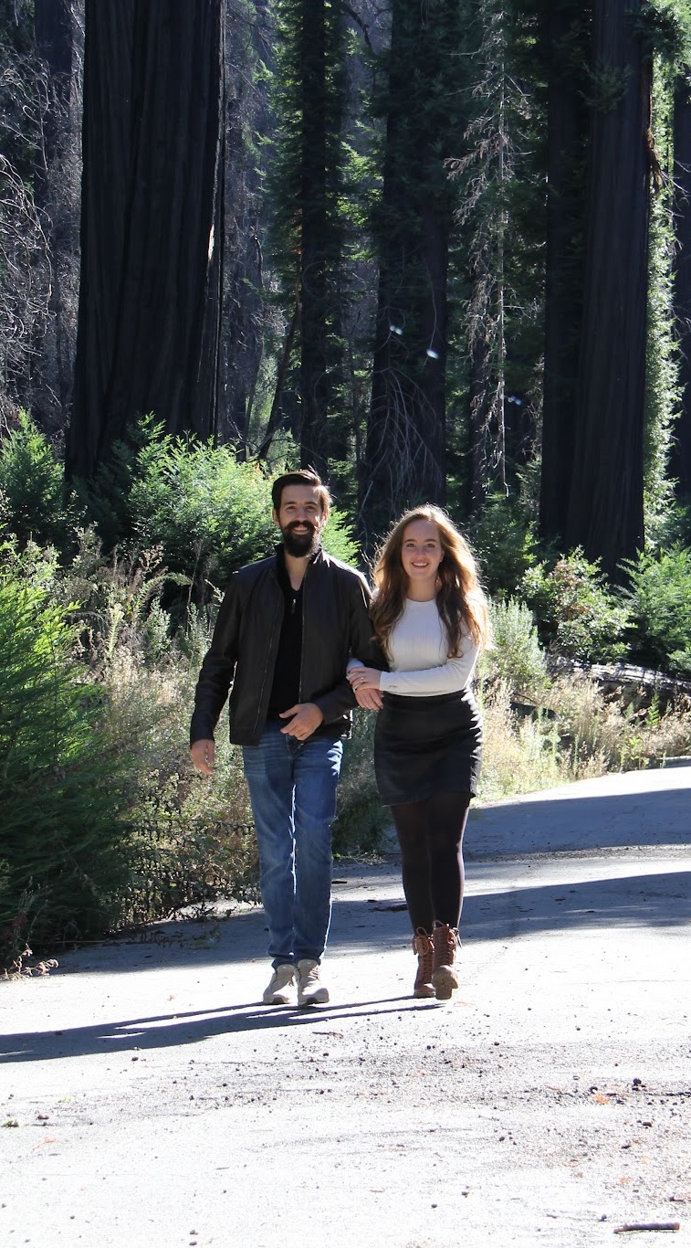 Natalie and Thomas walking down a road in the redwoods together, facing toward the camera and smiling.